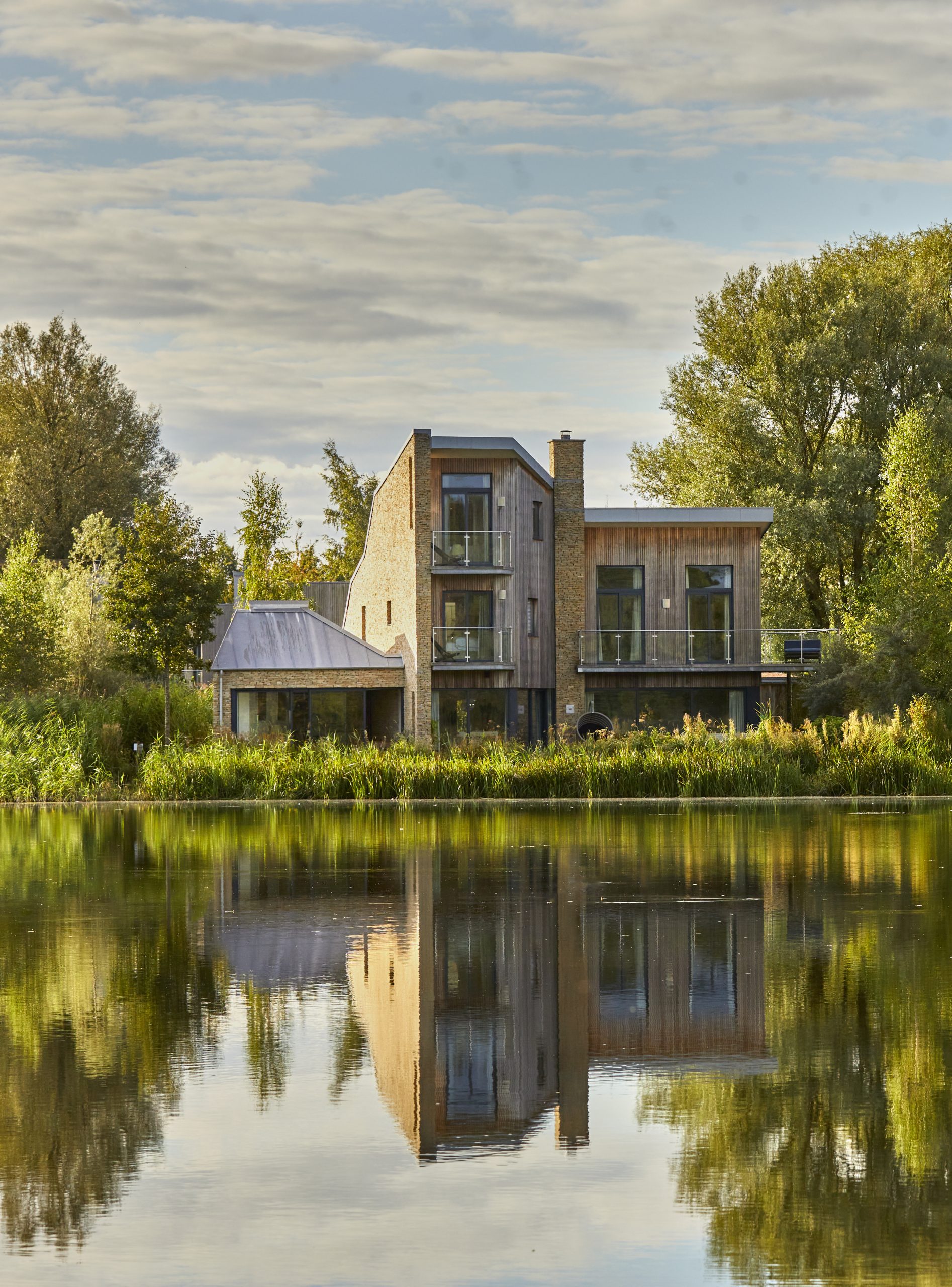 Modern two-story house with large windows and wood accents, surrounded by trees and greenery, reflected in a calm lake in the foreground.
